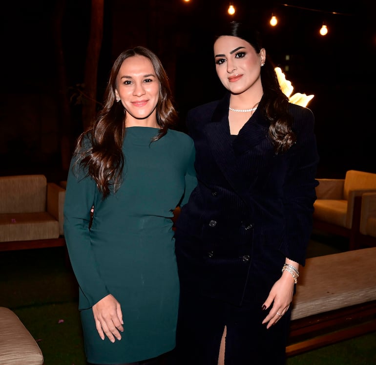 Dos mujeres sonrientes: una en vestido verde de manga larga y otra en abrigo oscuro, en ambiente acogedor y bien iluminado.