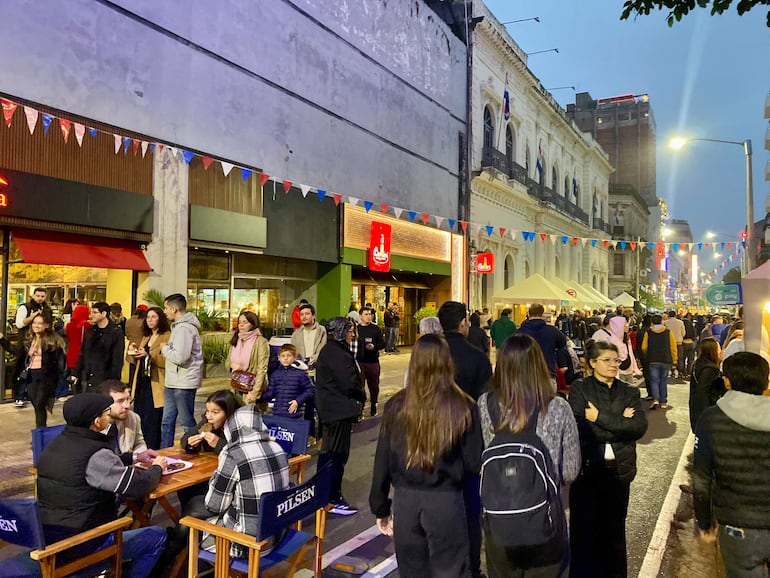 Dos personas sentadas en sillas conversan mientras otros asisten a un evento festivo con banderas rojas, azules y blancas.