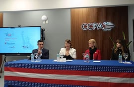 Carson Villalba en abrigo blanco habla en panel. Tres asistentes escuchan, uno en traje oscuro, otro en suéter rojo con gafas.