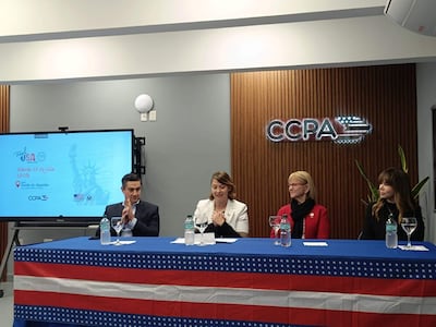 Carson Villalba en abrigo blanco habla en panel. Tres asistentes escuchan, uno en traje oscuro, otro en suéter rojo con gafas.