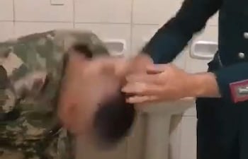 Militar vestido de uniforme inclinado hacia abajo, mientras otro superior con insignia lo golpea en un baño con azulejos.