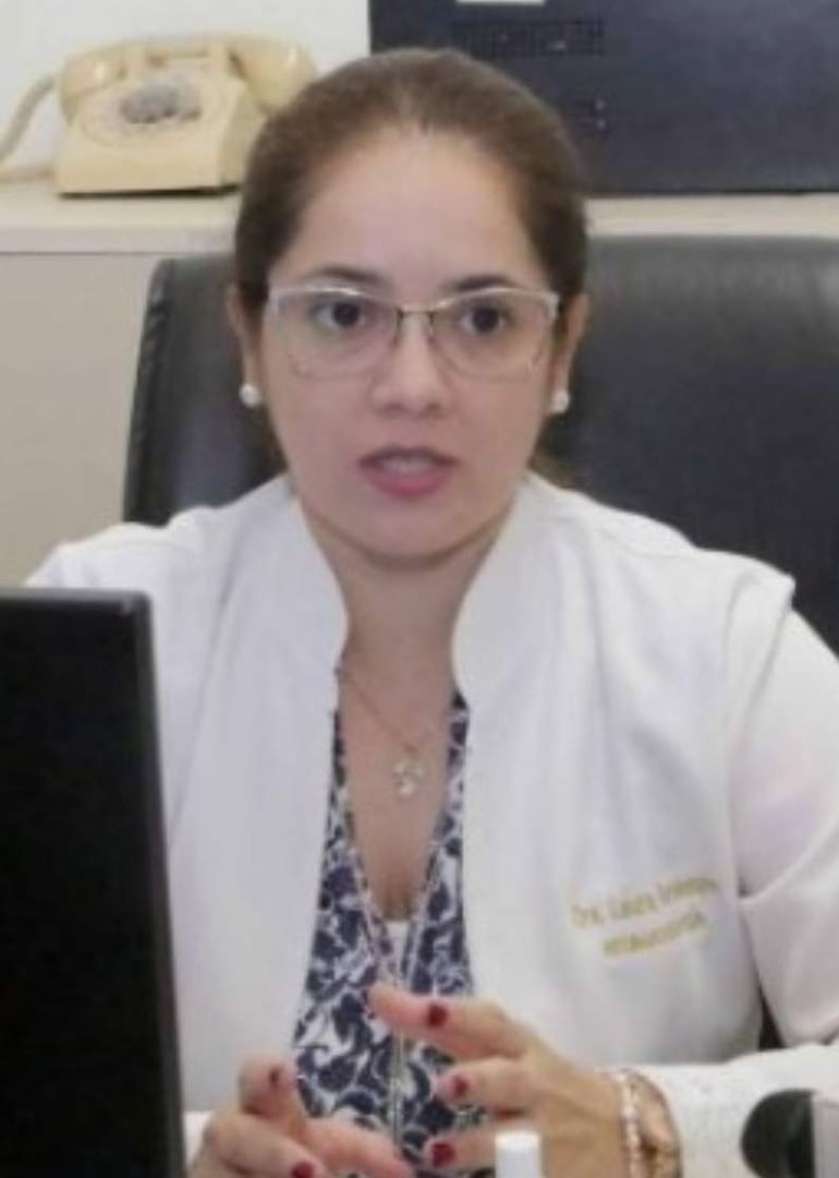 Dra. Laura Bordenave Gamarra con bata blanca, sentada en su despacho, gesticulando mientras habla, con teléfono antiguo y computadora a su alrededor.