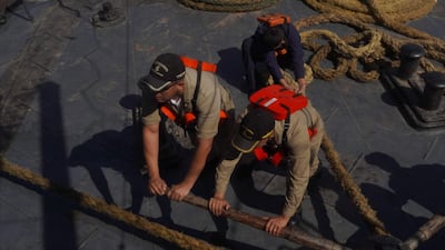 Tres hombres en chalecos salvavidas naranjas trabajando en un barco, sosteniendo cuerdas y palos de madera bajo los primeros rayos del sol.