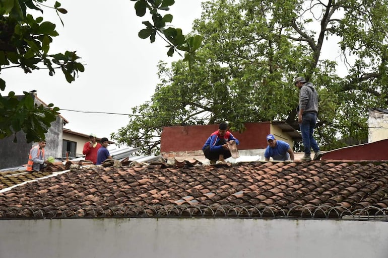 Cinco hombres en un techo de tejas, uno con chaleco naranja supervisa, mientras los otros sostienen tejas y trabajan. Entorno natural al fondo.