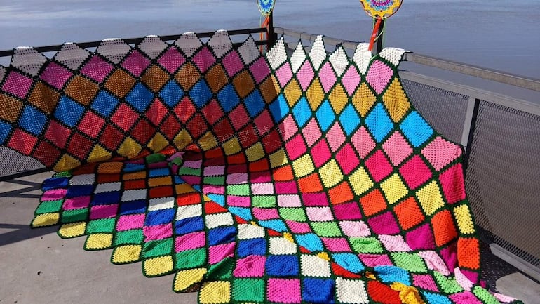 Tejido colorido de rombos en rosa, azul, amarillo y verde, colocado sobre una barandilla metálica en un entorno festivo.