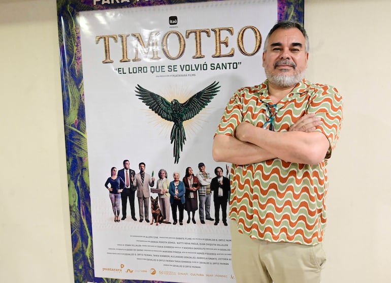 Hombre de pie junto a un cartel colorido de 'Timoteo', vistiendo camiseta marrón y verde, con brazos cruzados y expresión tranquila.