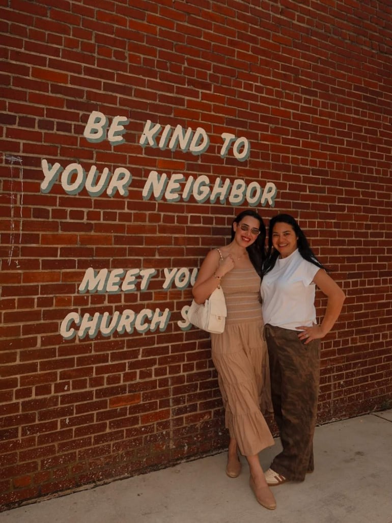 Alexia Notto con blusa de tirantes clara y Sonia Rejala en camiseta blanca con pantalones camuflados, sonrientes ante una pared de ladrillos.