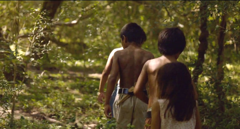 Tres niños descalzos caminan con la espalda hacia la cámara en un bosque frondoso, dos varones y una niña con cabello suelto.