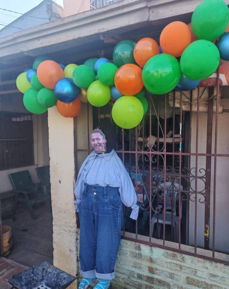Muñeco con rostro sonriente colgado sobre sillas verdes, rodeado de globos coloridos en ambiente festivo.