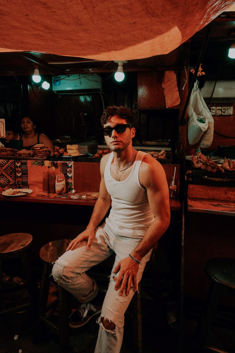 Acho Laterza vestido con camiseta blanca y gafas de sol, sentado en un puesto de comida con ambiente comunitario.