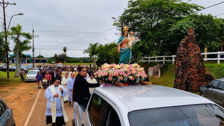 Sacerdote con vestimenta blanca lidera una procesión con la Virgen María Auxiliadora, rodeado de fieles y vehículos en un entorno religioso.
