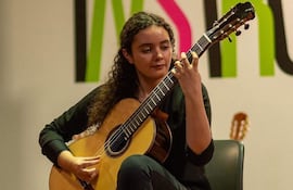 Mujer de cabello rizado y camiseta negra toca guitarra clásica en un ambiente artístico con una pared colorida al fondo.