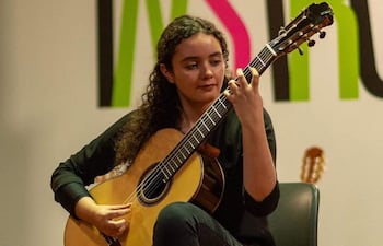 Mujer de cabello rizado y camiseta negra toca guitarra clásica en un ambiente artístico con una pared colorida al fondo.