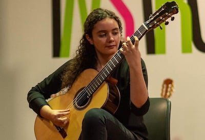 Mujer de cabello rizado y camiseta negra toca guitarra clásica en un ambiente artístico con una pared colorida al fondo.