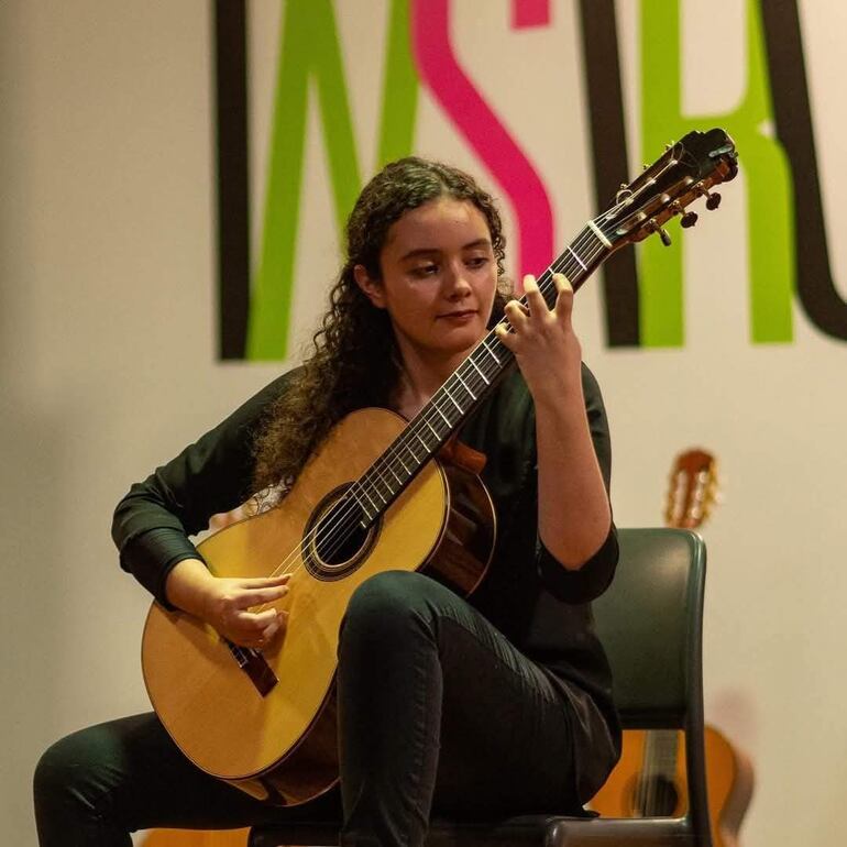 María Paz Cubells, con cabello rizado y camisa negra, toca la guitarra en un escenario con fondo colorido.