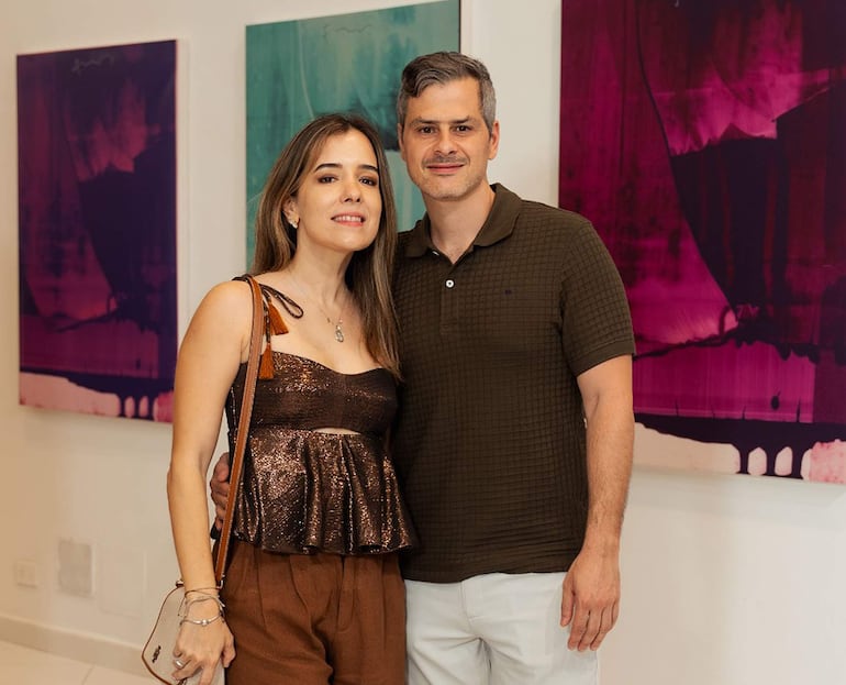 Mujer con blusa marrón y hombre en camisa oliva, ambos sonríen en una galería decorada con arte en tonos púrpuras y verdes.