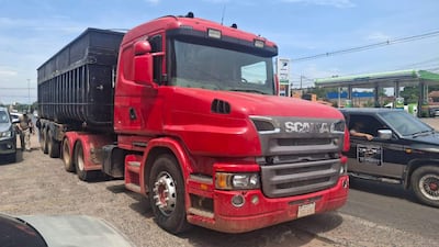 Camión rojo Scania detenido, con una persona de pie a su izquierda, en un entorno soleado y urbano.