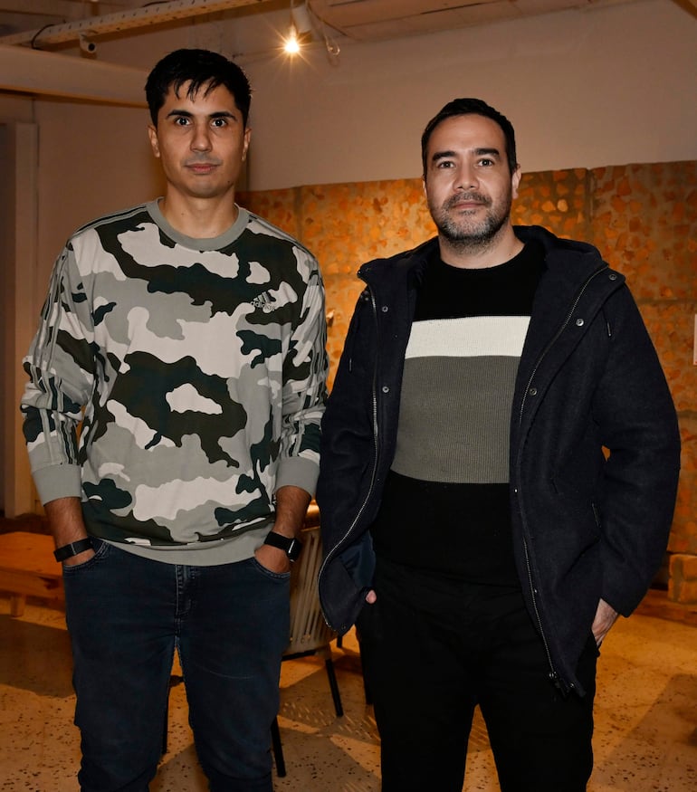 Dos hombres, uno con camiseta de camuflaje y otro con abrigo oscuro, posan en un entorno moderno y artístico.