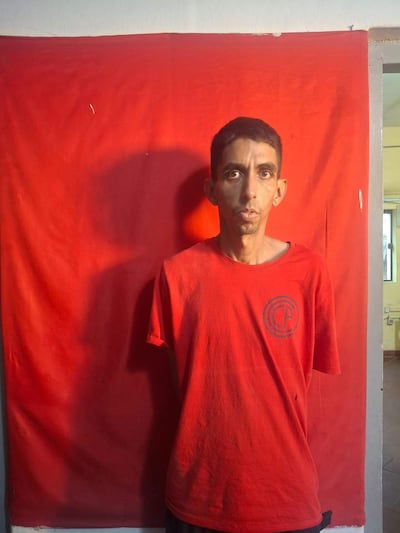 Hombre de pie con camiseta roja y expresión neutral, frente a fondo rojo en ambiente cerrado.