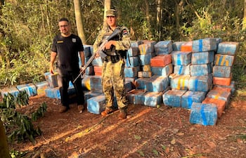 Hombre en camiseta negra conversando con otro en uniforme camuflado, rodeados de paquetes azules y naranjas en un área boscosa.