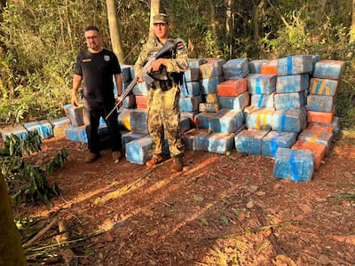 Hombre en camiseta negra conversando con otro en uniforme camuflado, rodeados de paquetes azules y naranjas en un área boscosa.
