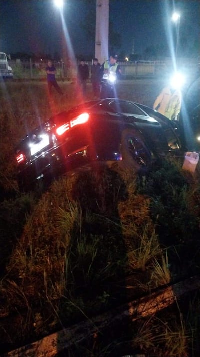 Automóvil negro accidentado en terreno de hierba, con policías y curiosos observando la escena iluminada por farolas.