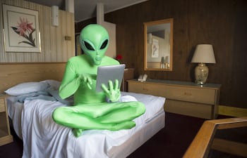 Extraterrestre verde sentado con una tablet en la cama en una habitación anticuada, imagen ilustrativa.