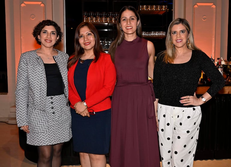 Cuatro mujeres sonriendo en un salón elegantemente decorado, vestidas con ropa formal y posando juntas.