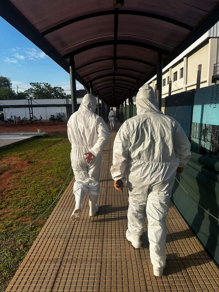 Tres personas con trajes de protección blanca caminan por un pasillo cubierto, mientras áreas verdes y construcciones se ven a su alrededor.