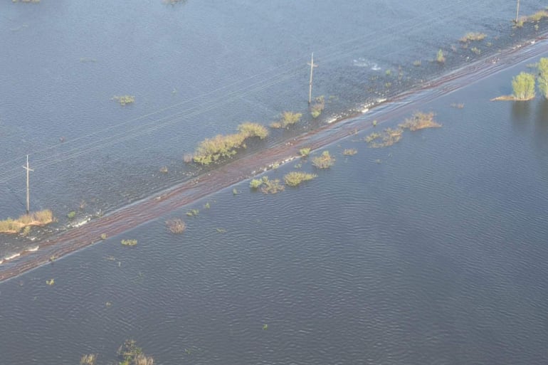 Vista aérea de una carretera parcialmente sumergida rodeada de agua y vegetación, un poste eléctrico visible a la izquierda.