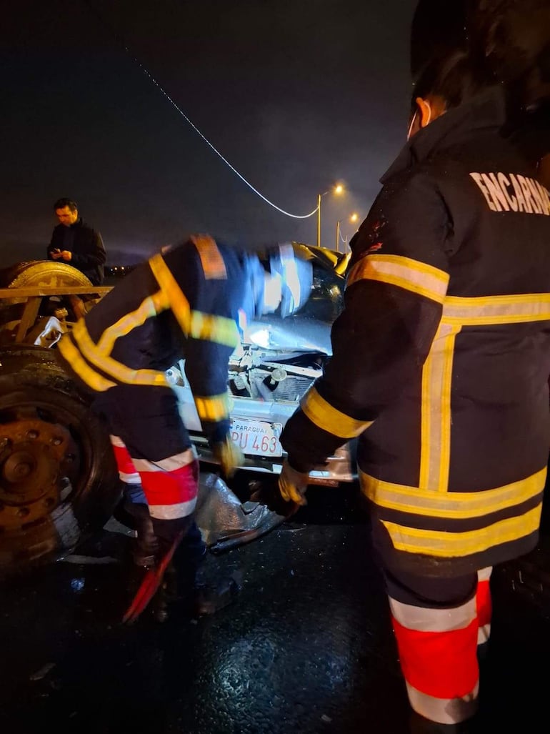 Bomberos de uniforme negro y rojo ayudan a un herido en un vehículo destrozado bajo luz de faroles en la noche.