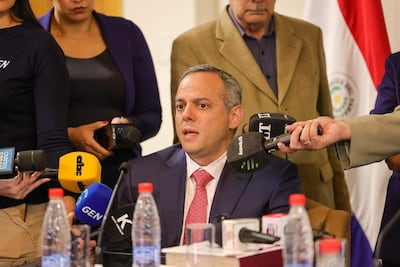 Camilo Benítez Aldana habla en conferencia, vestido de traje oscuro con corbata rosada, rodeado de micrófonos y asistentes atentos.