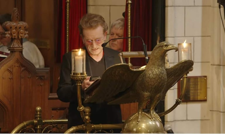 Hombre de pie en podio con forma de águila, leyendo en un ambiente ceremonial con velas.