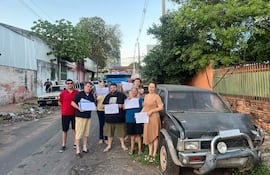 Grupo de seis personas, incluyendo hombres y mujeres, sosteniendo documentos en una calle con un auto en mal estado al fondo.