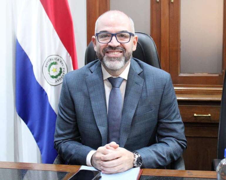 Enrique Javier Berni Brítez, hombre de traje negro y corbata, sonríe en su escritorio con fondo de bandera paraguaya.