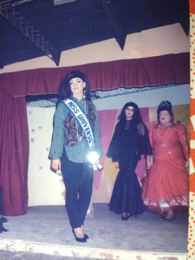 Competidora Miss Universo con banda en blusa estampada y pantalones oscuros, acompañada por dos personas en traje y vestido rojo.