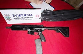 Fusil Colt AR-15 sobre mesa roja con cartulina de 'EVIDENCIA ANTINARCÓTICOS' y una mano sosteniendo un cuaderno.
