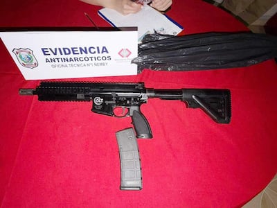 Fusil Colt AR-15 sobre mesa roja con cartulina de 'EVIDENCIA ANTINARCÓTICOS' y una mano sosteniendo un cuaderno.
