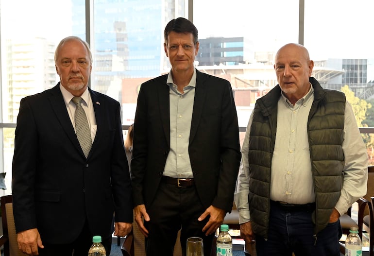 Tres hombres de pie, uno en traje oscuro, otro en blazer negro y el tercero en chaleco gris, posan en sala con grandes ventanales.