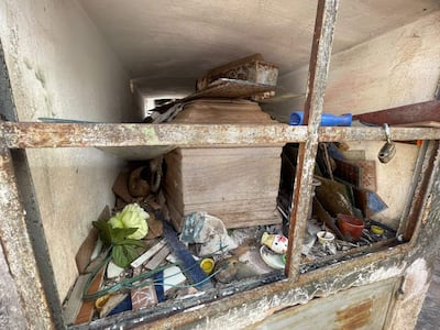 Estructura cerrada en cementerio con rejas oxidadas. Dentro, cajas y objetos en desorden, con adorno floral artificial.
