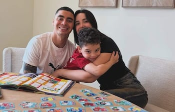 Miguel Almirón sonriente sosteniendo álbum, mujer abrazando a niño con camiseta roja, tarjetas de fútbol en la mesa.