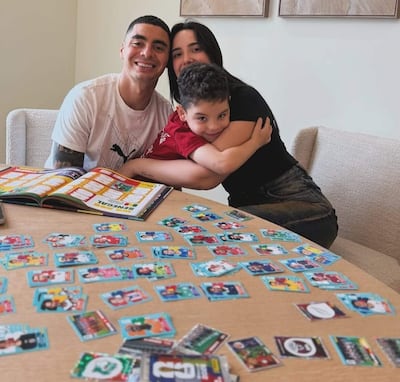 Miguel Almirón sonriente sosteniendo álbum, mujer abrazando a niño con camiseta roja, tarjetas de fútbol en la mesa.