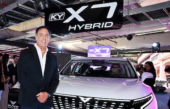 Hombre vestido de traje negro junto a SUV blanca 'KY X7 HYBRID'. Mujer con cámara detrás, capturando momentos del evento.