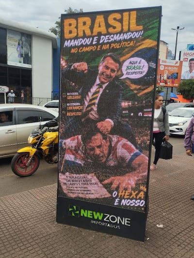 Cartel digital en área comercial, con figura masculina destacada y mensaje político sobre Brasil, rodeado de gente y vehículos.