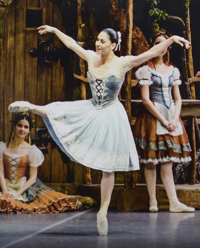 Bailarina principal en tutú azul claro, otra de rodillas en vestido marrón, y una más de pie con vestido colorido, todos en un ambiente teatral.