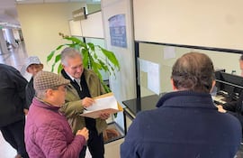 Dos hombres discuten documentos en un entorno administrativo; uno viste chaqueta verde y el otro lleva gorra de cuadros y abrigo burdeos.