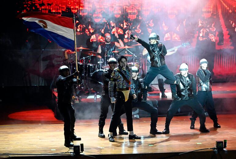Artistas en escenario con trajes brillantes y chaquetas negras, mientras uno sostiene una bandera de Paraguay en un entorno lleno de luces.
