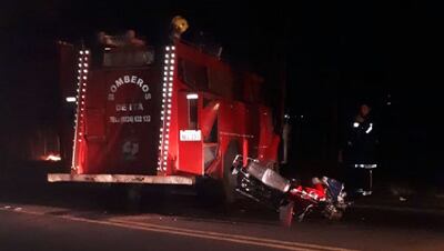 En la imagen se aprecia la motocicleta tumbada en el pavimento, tras impactar contra la parte lateral trasera del camión de bomberos. De fondo, el fuego.