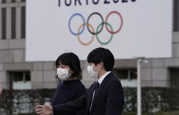 Peatones con tapabocas caminan cerca de un emblema de los JJ.OO., en Tokio. El evento sigue programado iniciar el 24 de julio.