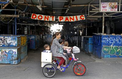 Una mujer monta una bicicleta con sus hijos frente a una pancarta colgada en la entrada del mercado Shuk HaCarmel (Caramelo) en la ciudad costera israelí de Tel Aviv.